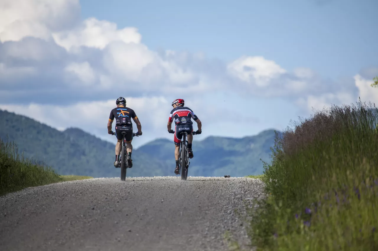 Two men ride their bikes on a gravel trail up a hill surrounded by tall grass. 
