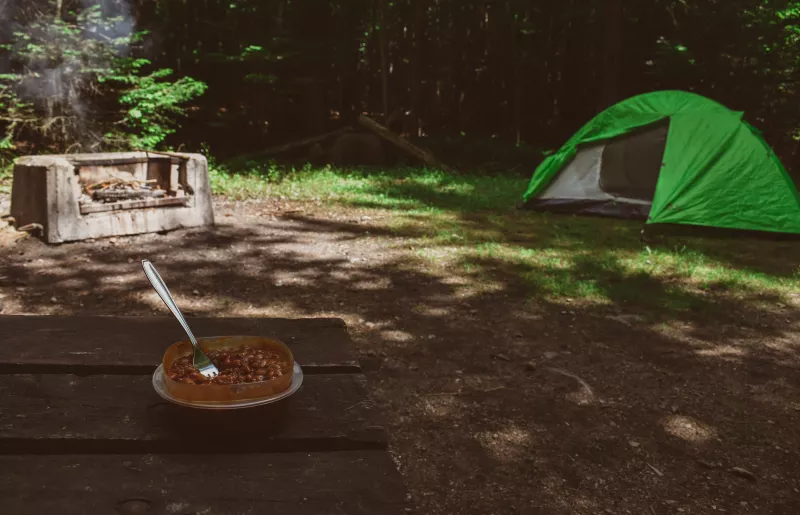 A campfire, a green tent, and a bowl of baked beans on a picnic table.