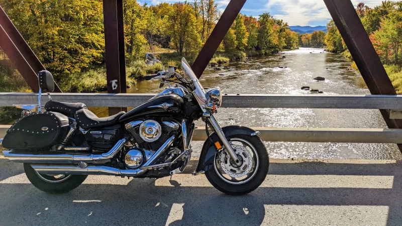 A motorcycle stands on a bridge overlooking a sparkling river. Fall foliage and mountains are in the background.