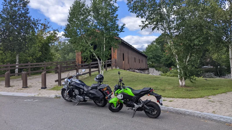 Two motorcycles sit parked in front of a grassy lawn and wooden covered bridge.
