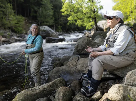 A woman stands in a river, fly fishing, while another sits on a riverside rock on a sunny day in a forest.