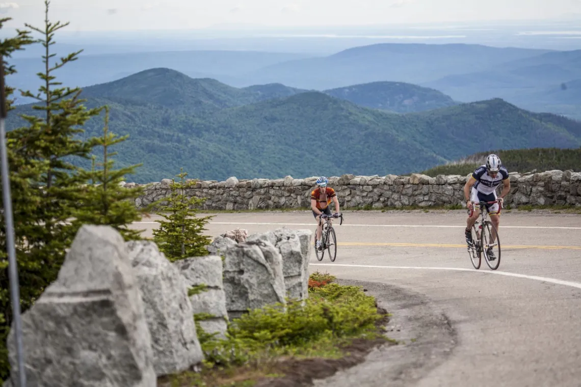 Two cyclists ride up Whiteface Mountain Memorial Highway with the mountains in the background.