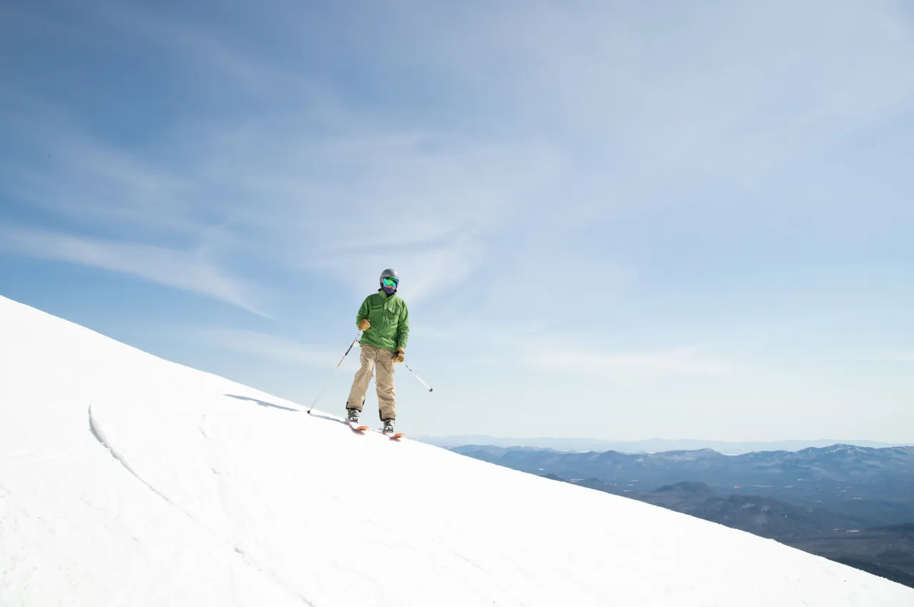 Skier in green jacket stands on slope with blue sky and mountains behind