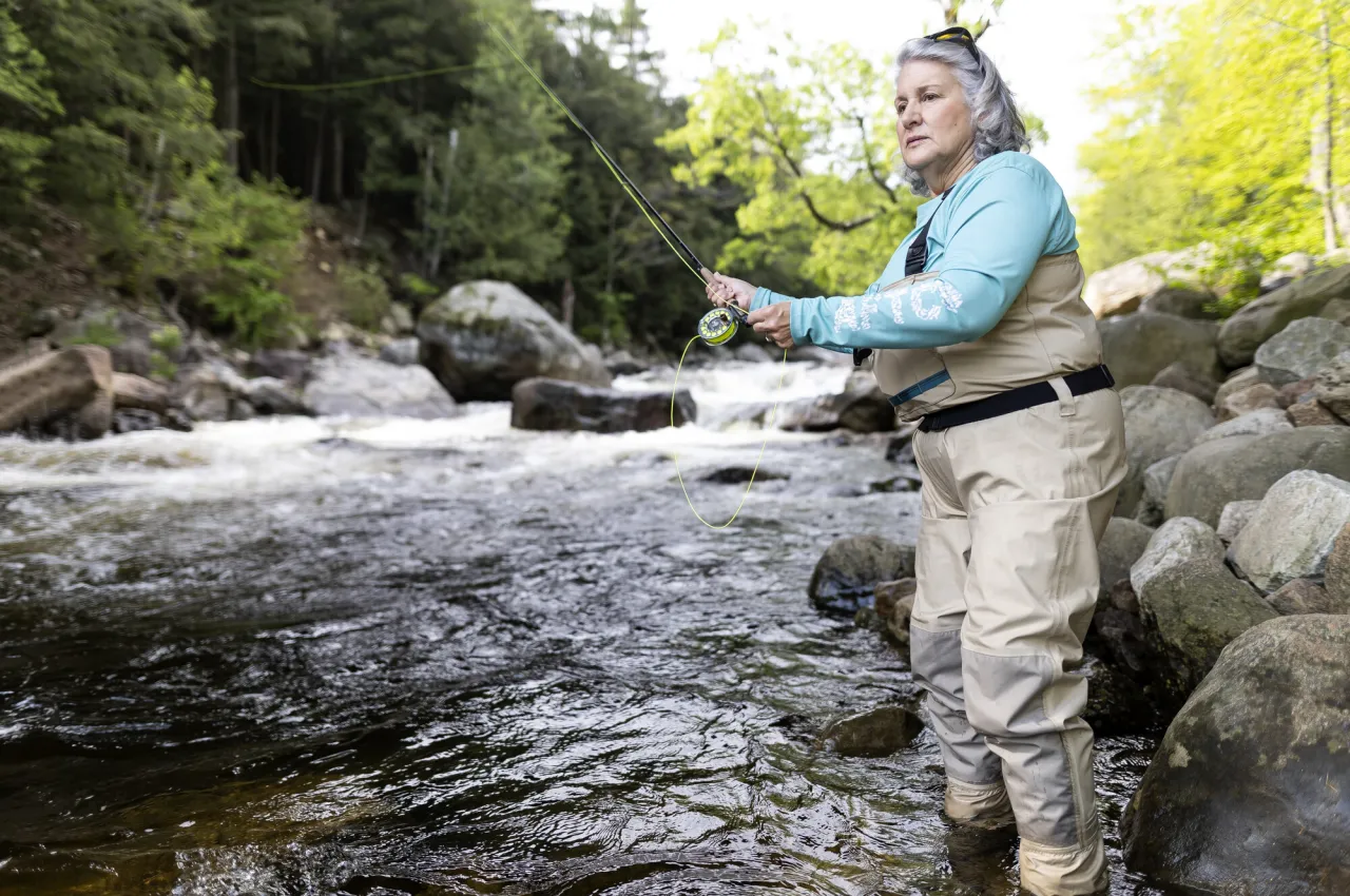 A woman in waders casts her line into a river on a sunny day.