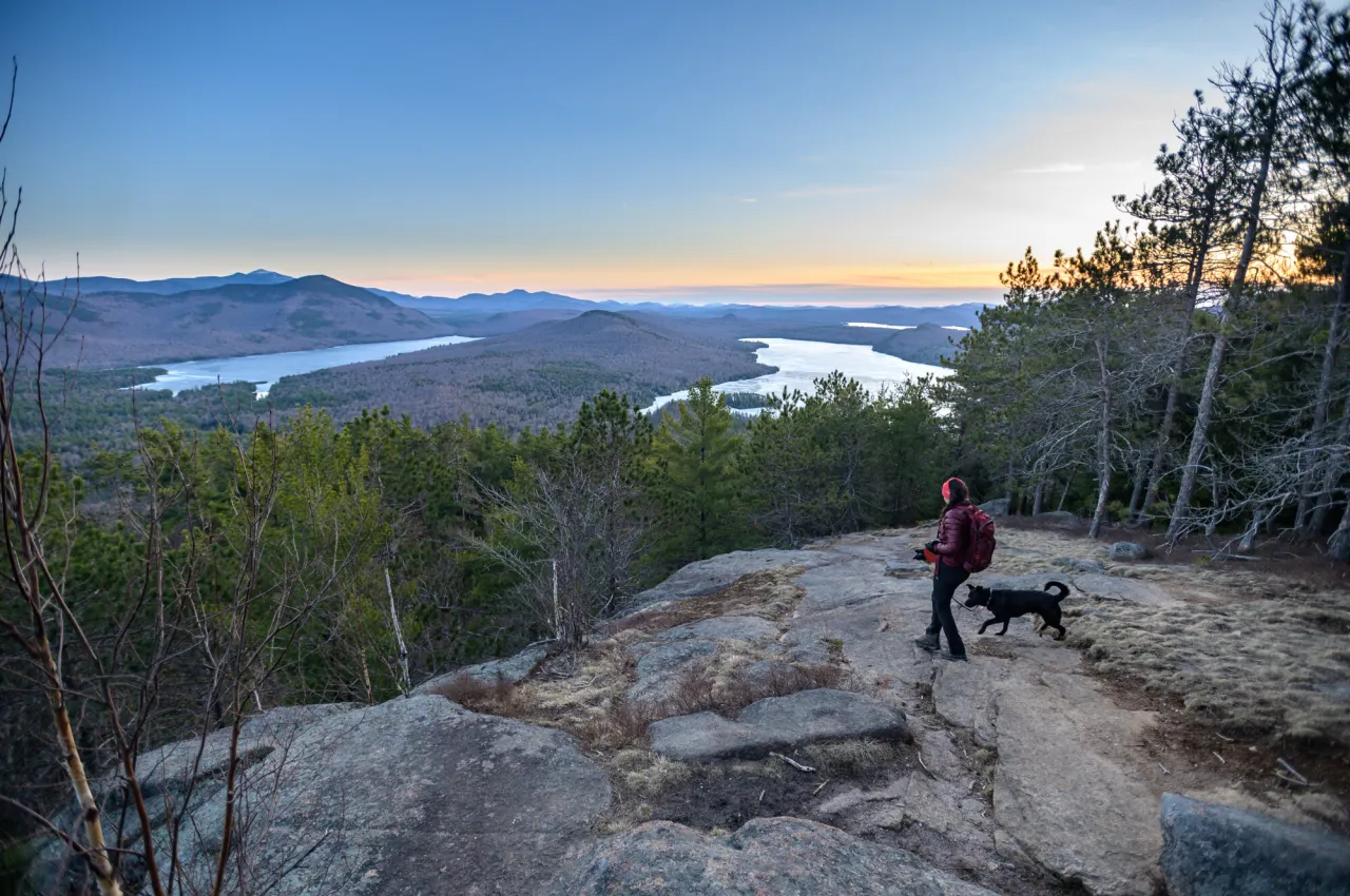 A woman and a black dog on a rocky summit overlooking forest and a distant lake at dusk.