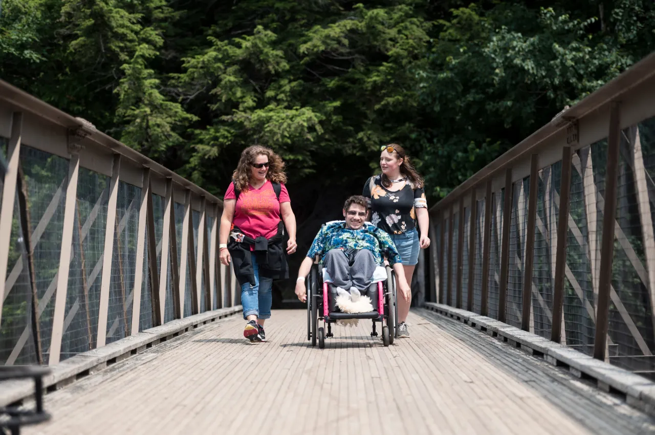 A person in a wheelchair is accompanied by a couple other people crossing a wooden bridge.
