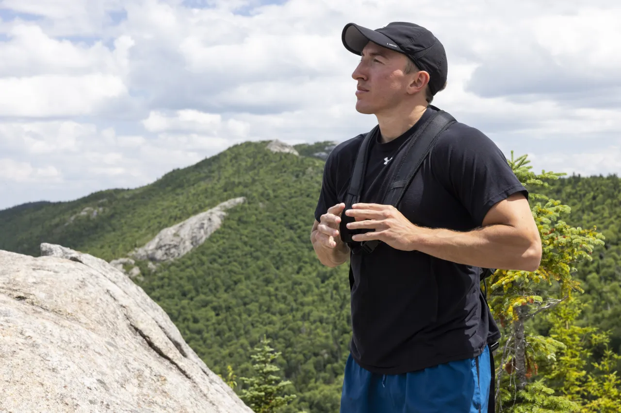 A male hiker wearing a tshirt and baseball cap looks upward on a rocky peak, with a forested mountain in the background.