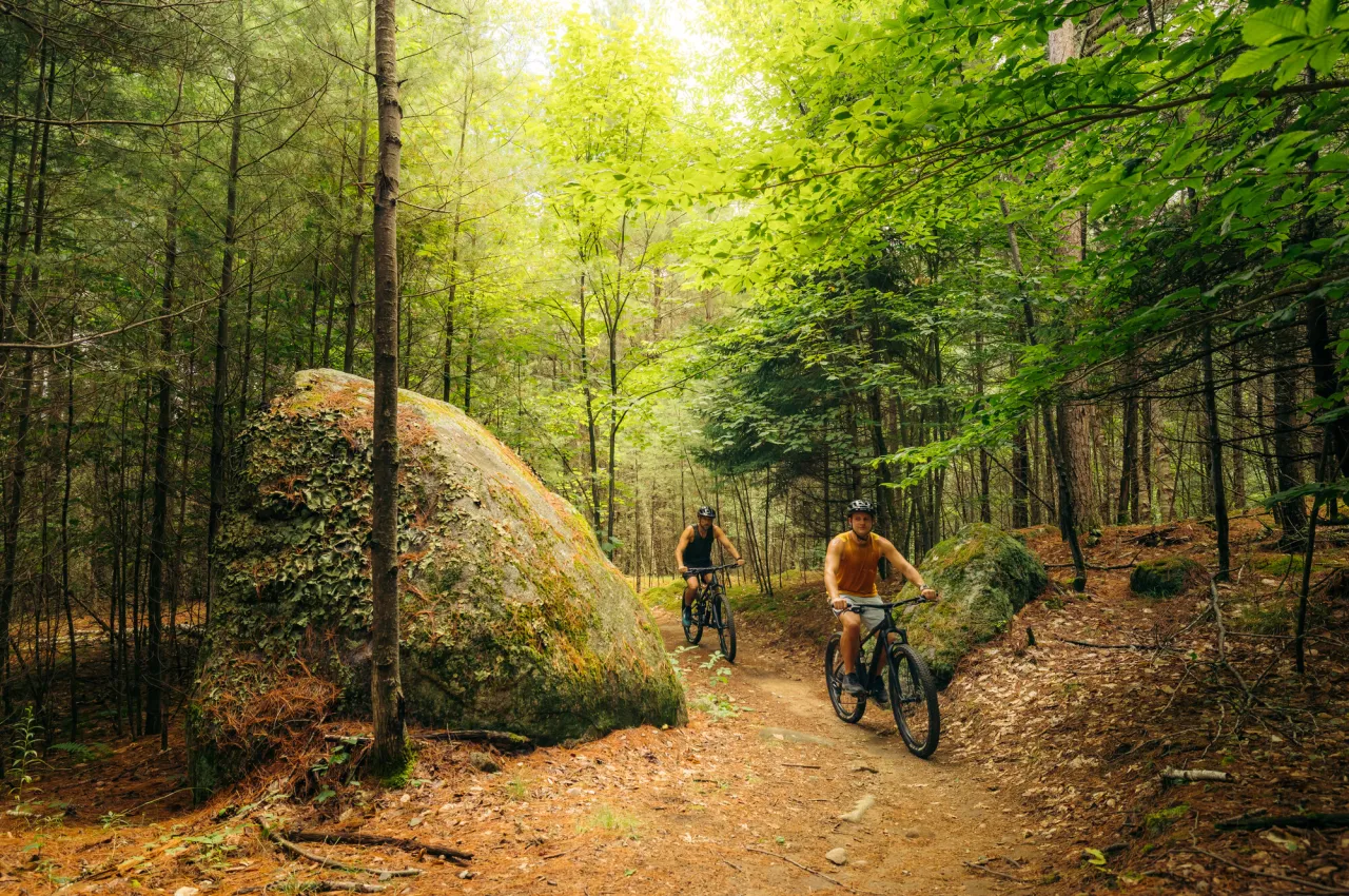 Two people on mountain bikes ride past large boulder on forested trail