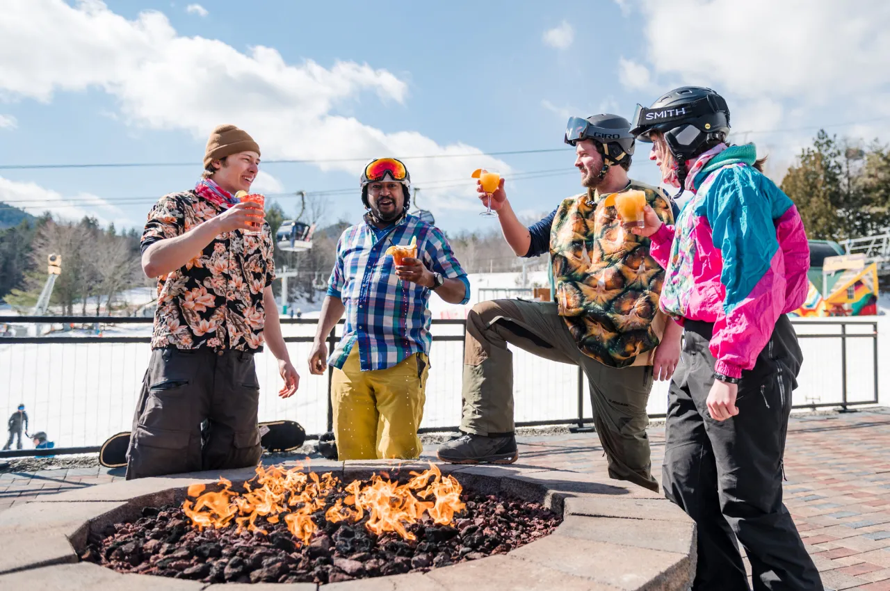 Four people in ski gear and costumes stand around outdoor fire holding drinks
