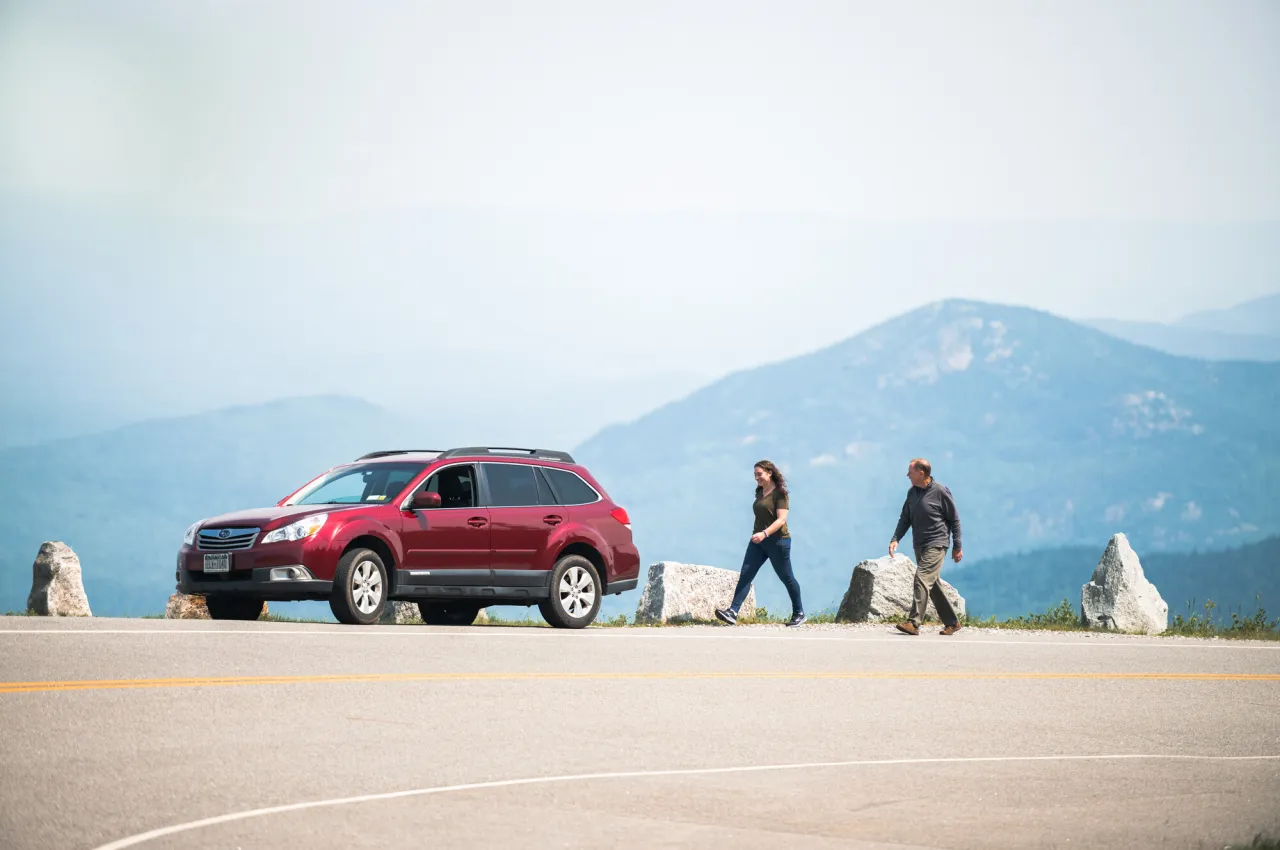Man and woman walk along road back toward red car with mountain views in background