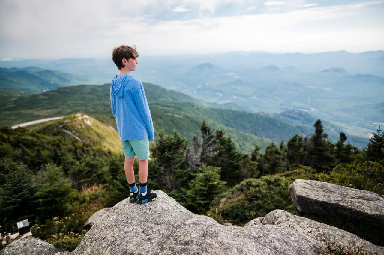 Boy stands on rock on mountain summit looking out at view