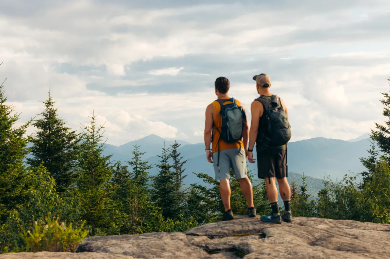 Two men stand side by side on mountain top looking out at view with backpacks on