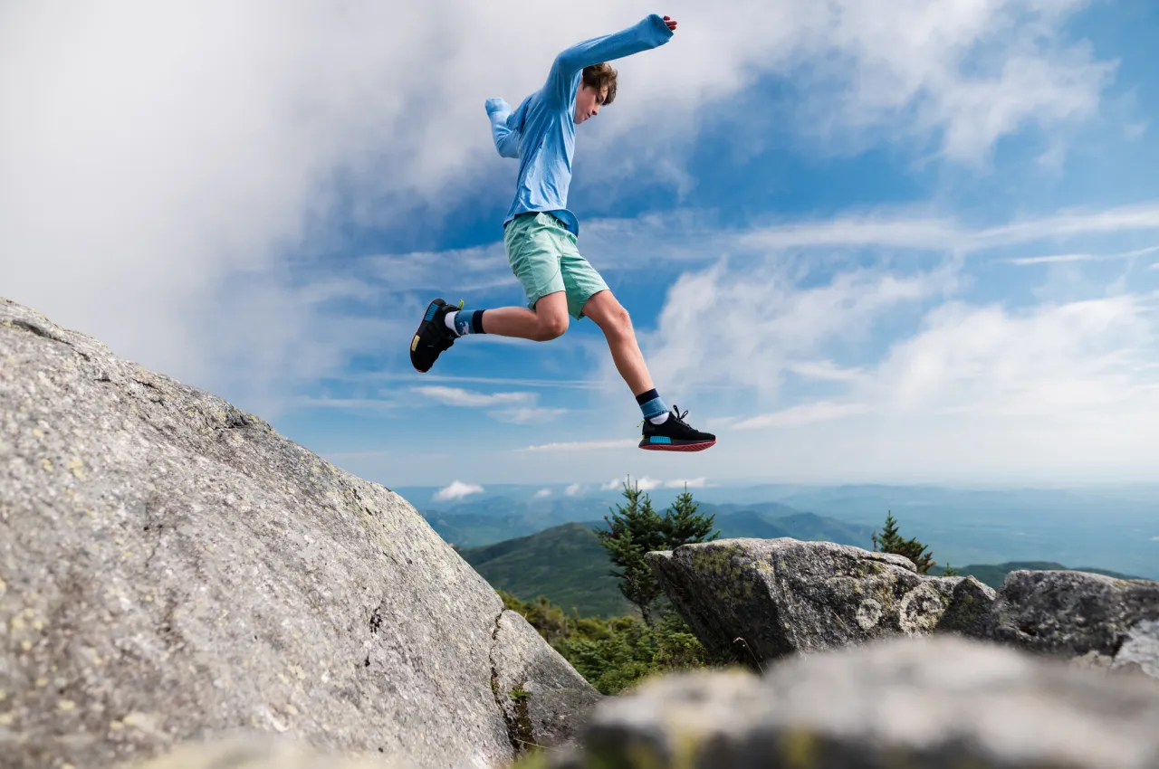 Adolescent boy in shorts and blue long sleeve shirt jumps from high rock to low rock on mountain summit