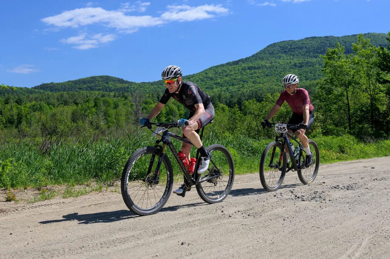 Two mountain bikers ride single file on gravel road in summer with green field and mountain view behind them