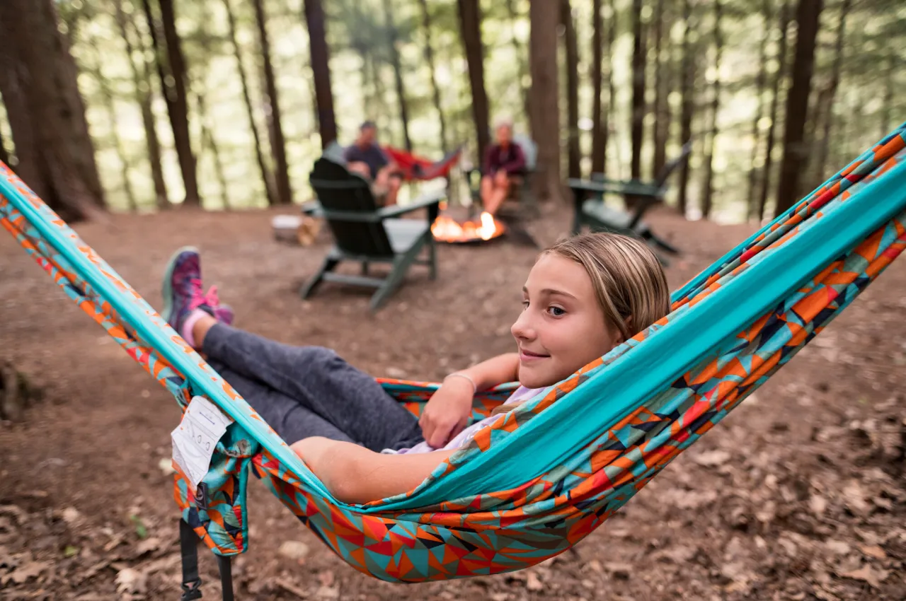 Adolescent girl lies in colorful hammock while parents and brother sit around fire in Adirondack chairs behind her