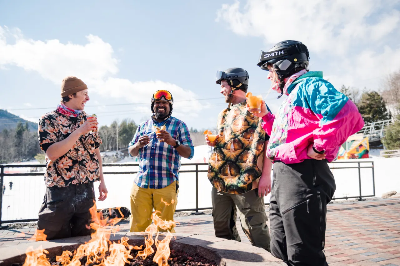 Four friends in ski gear and hawaiian clothes drink by fire on deck at ski resort