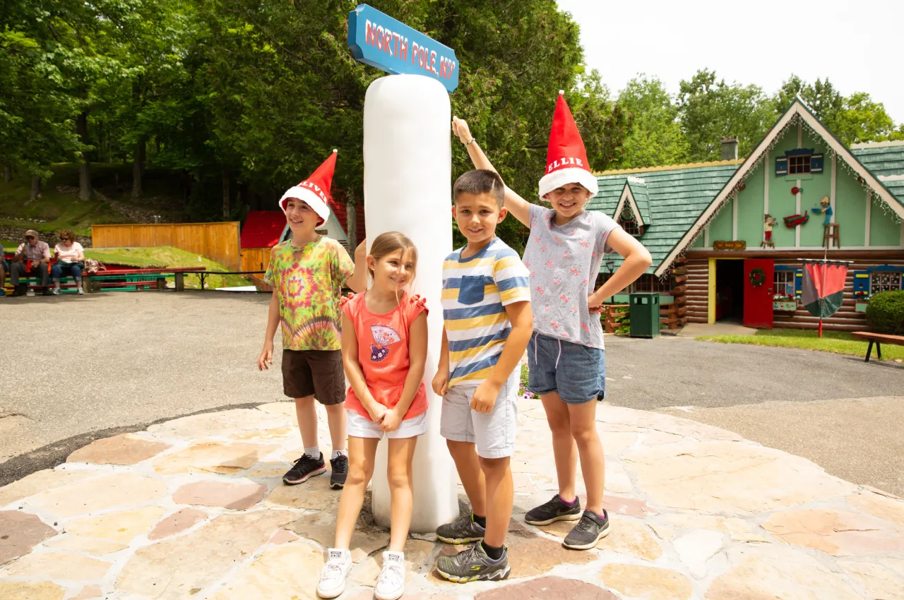 Four kids stand next to North Pole pole, two with red elf hats on, on summer day