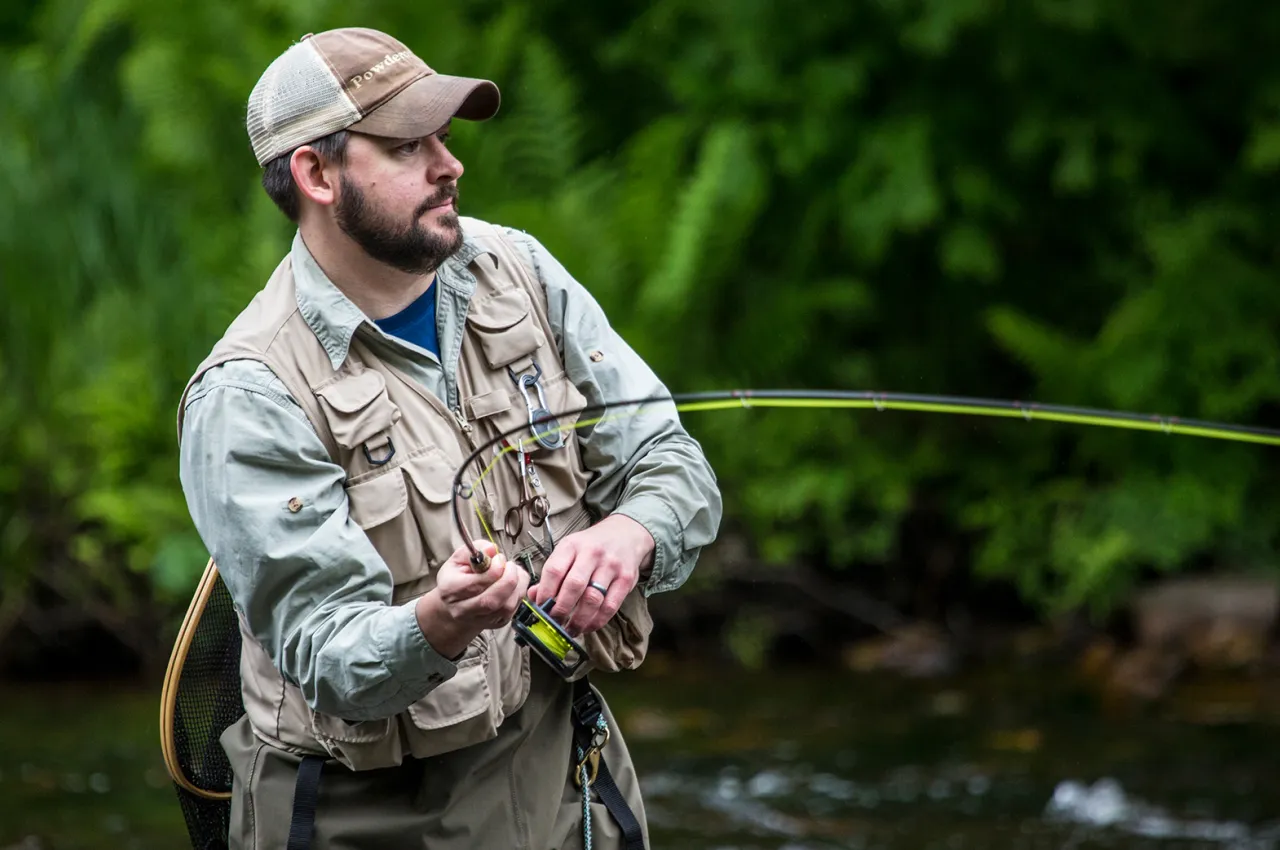 A man in fishing shirt, vest, and baseball cap holds a bent fly rod against a deep green background.