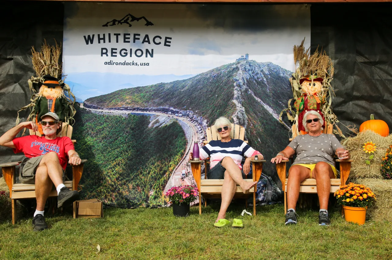 Three people sit in front of a whiteface region sign at a festival.