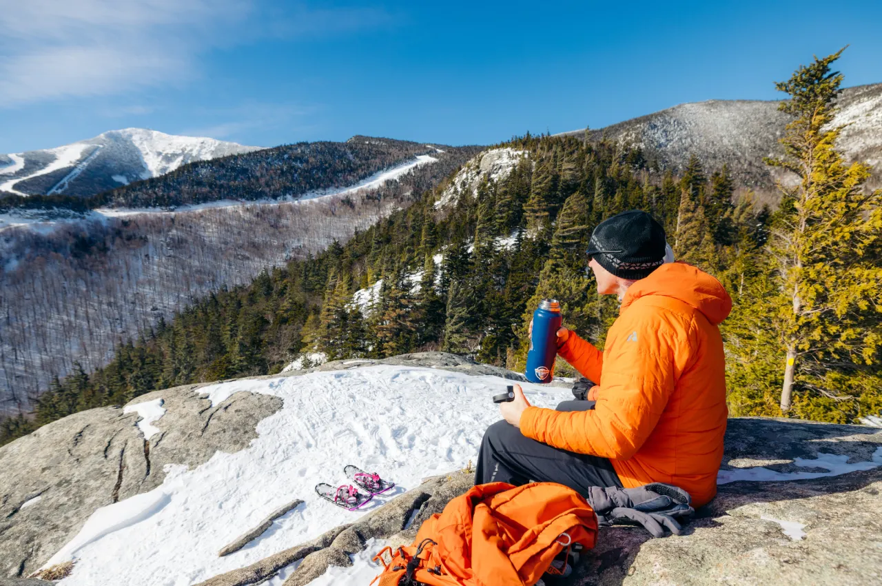 A man drinks from a thermos on top of a mountain in winter. 