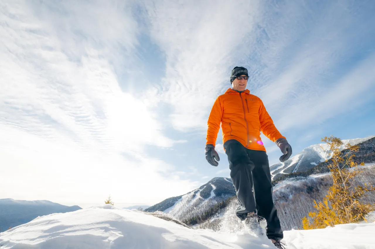 A man in an orange jacket walks through snow on a clear day. 