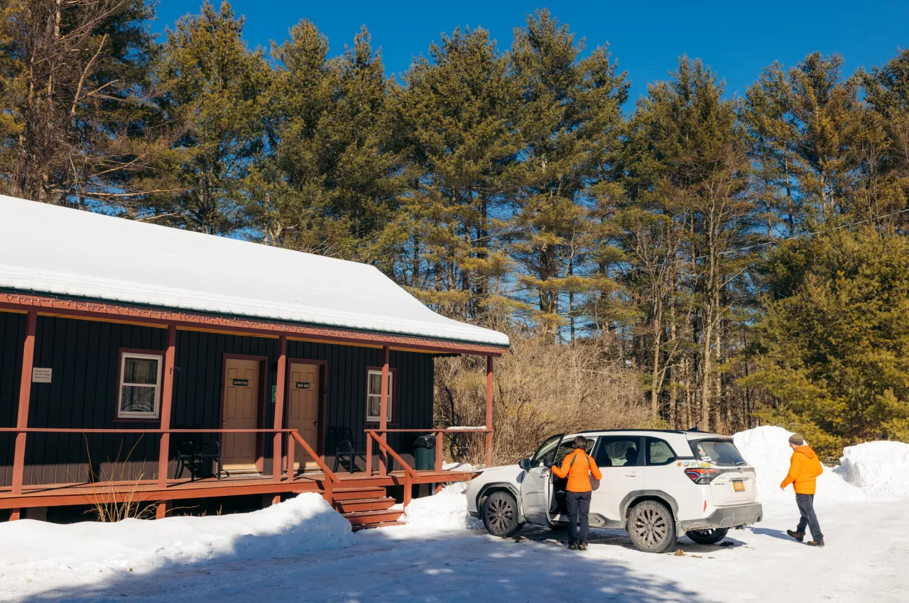 Two people unload a white car in front of a brown cottage in winter. 