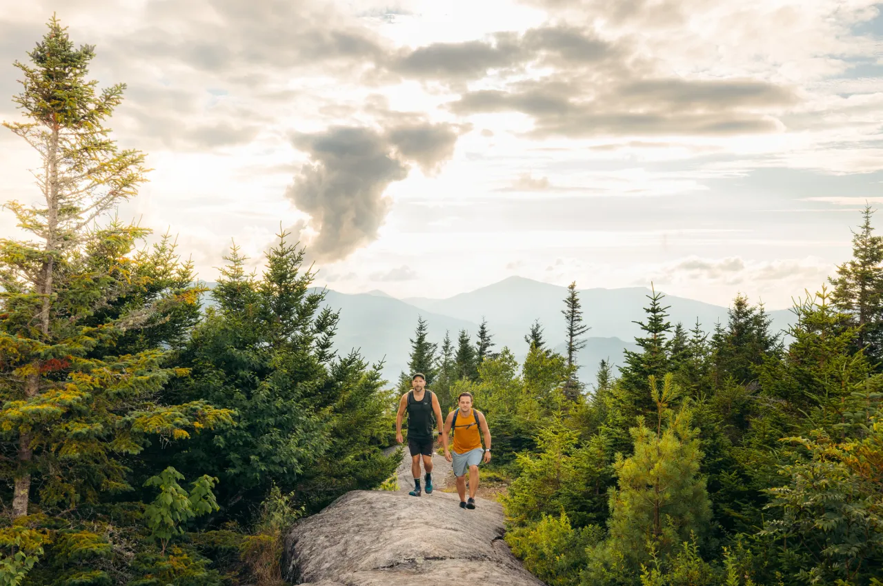 Two men ascend a rock slab trail between mostly pine trees on a mountain with other faded mountains visible in the background