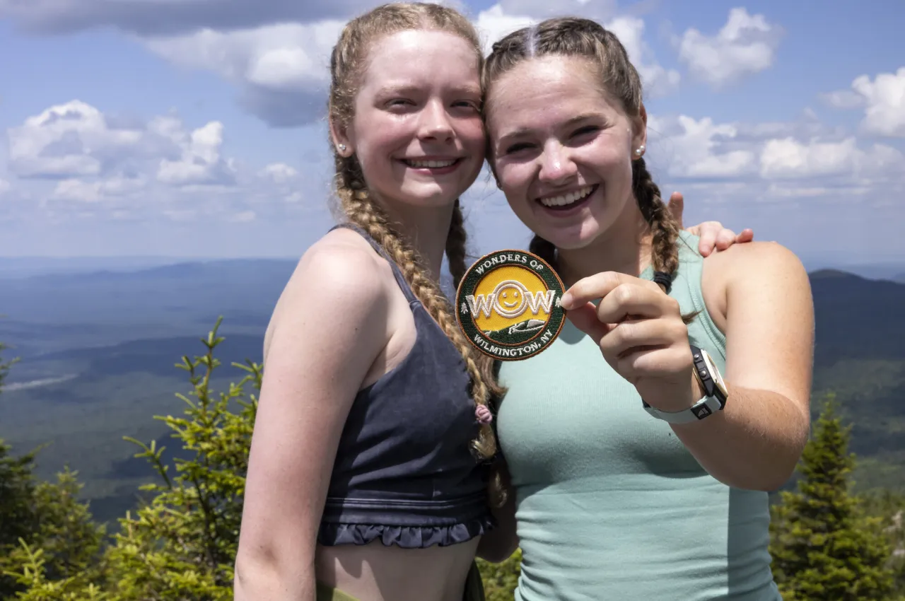 Two young woman with long hair in braids stand on mountain top smiling, one holding out a patch