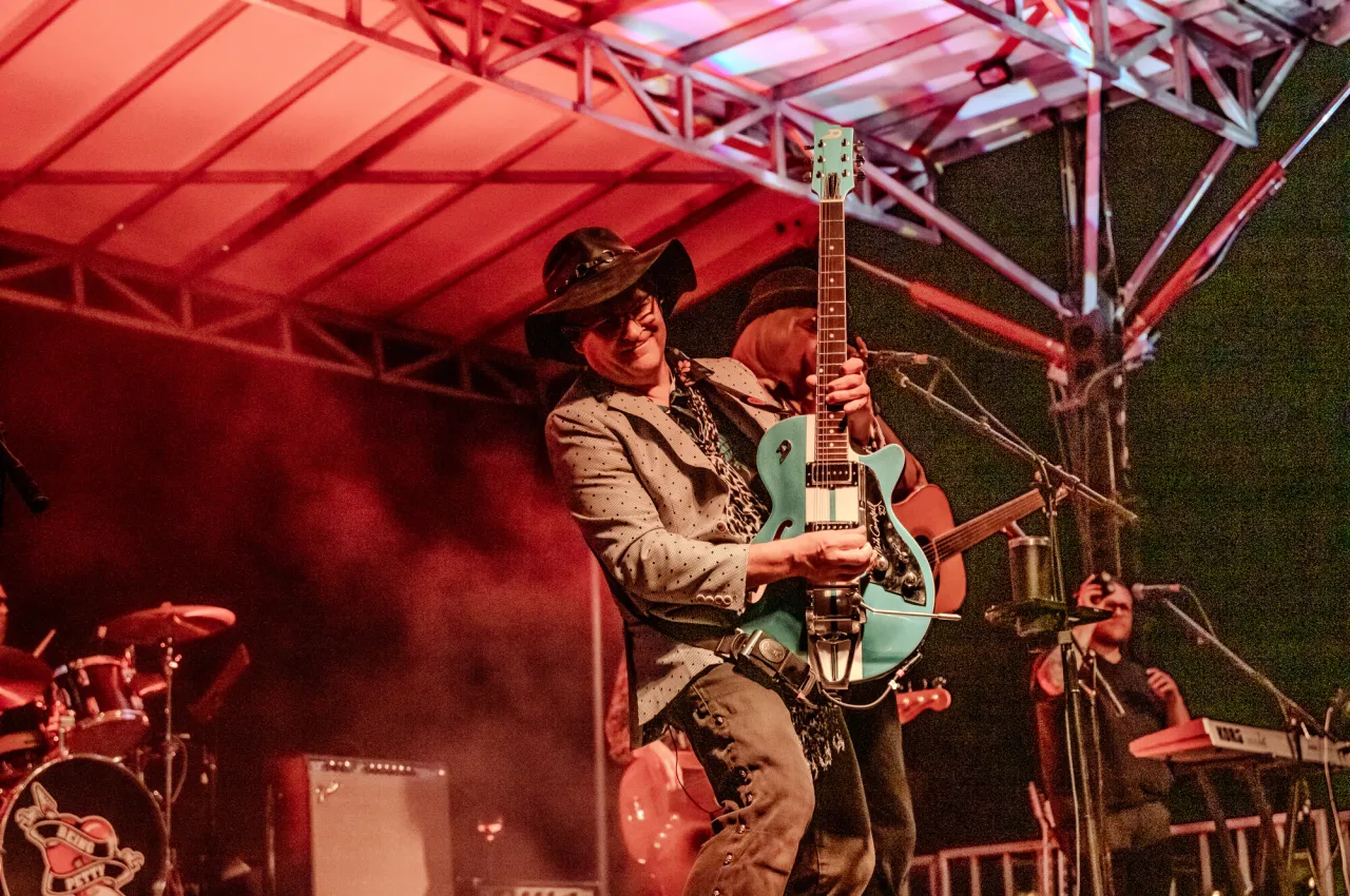 Man in cowboy hat on red-lit stage holds teal electric guitar upright as he plays