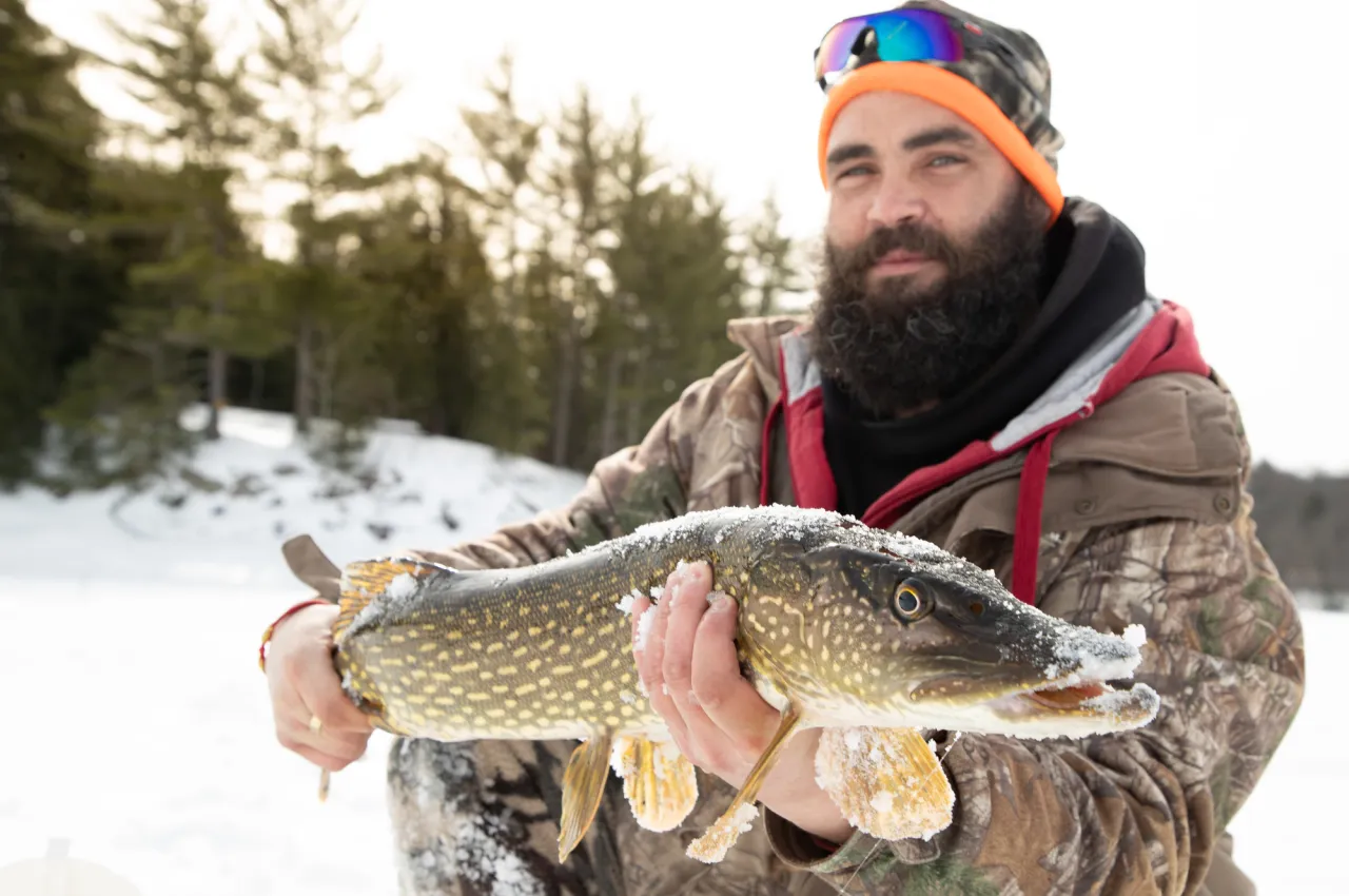 A man with a beard holds a fish on a frozen lake in winter.