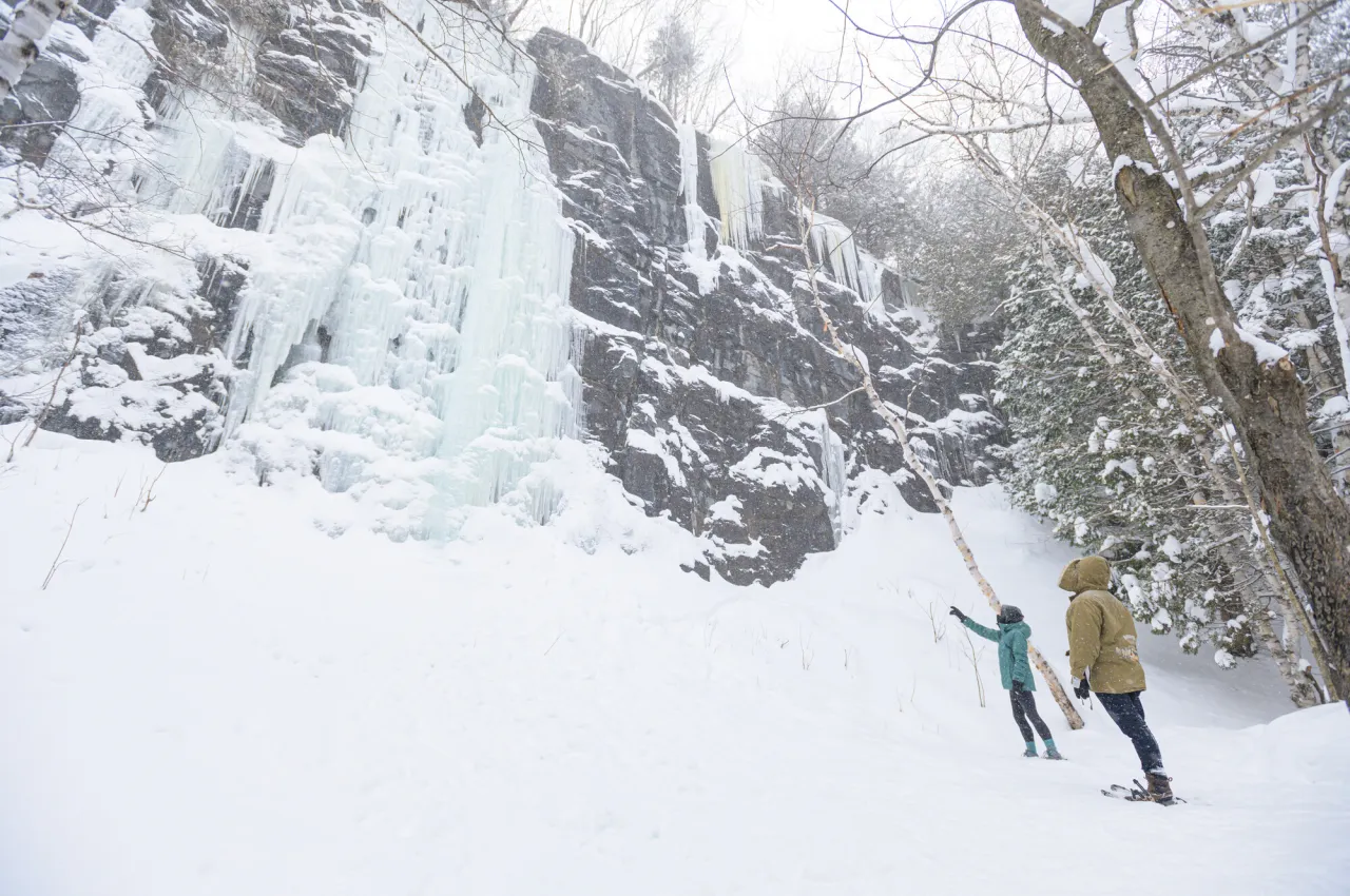 A man and woman observe an ice formation cascading down rocks on a winter trail. 