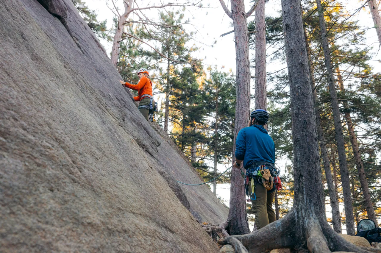 Man wearing climbing gear and helmet belays for man climbing on diagonal rock face in front of him surrounded by pine trees