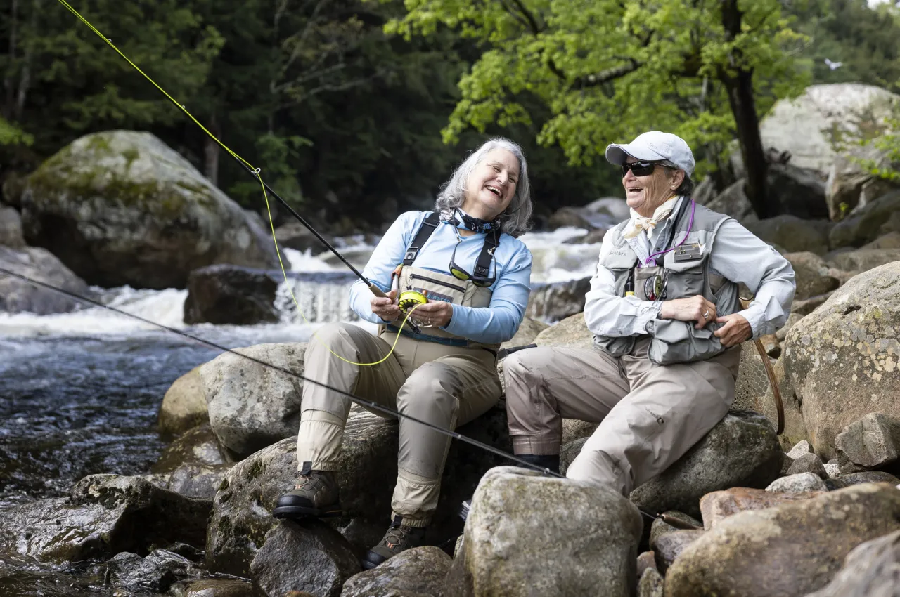 Two senior women in waders sit on rocks casting fishing lines into river and laughing