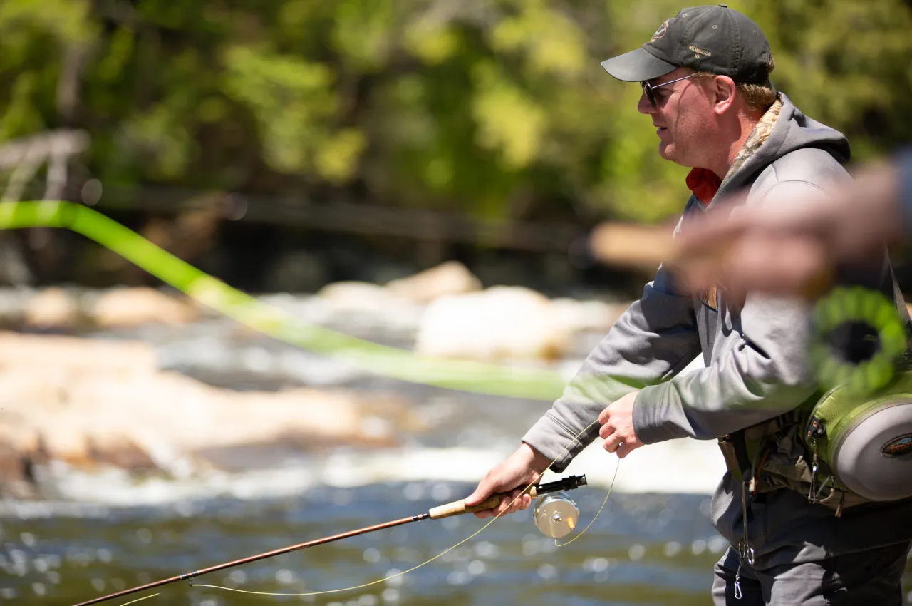 A man casts a fly fishing line on a sunny day. Water, rocks, and trees are blurred in the background.