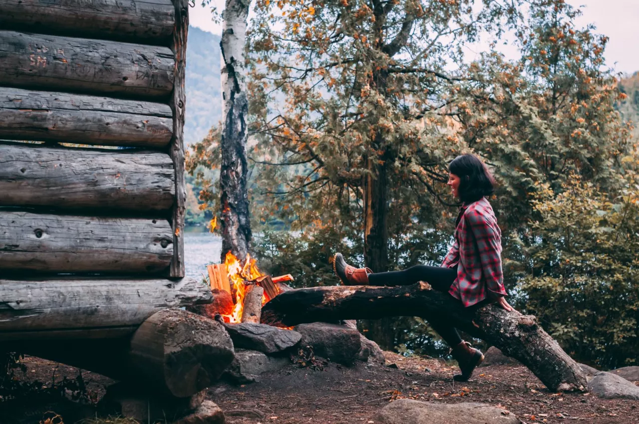 A woman sits on a log near a lean-to, warming herself next to a roaring fire
