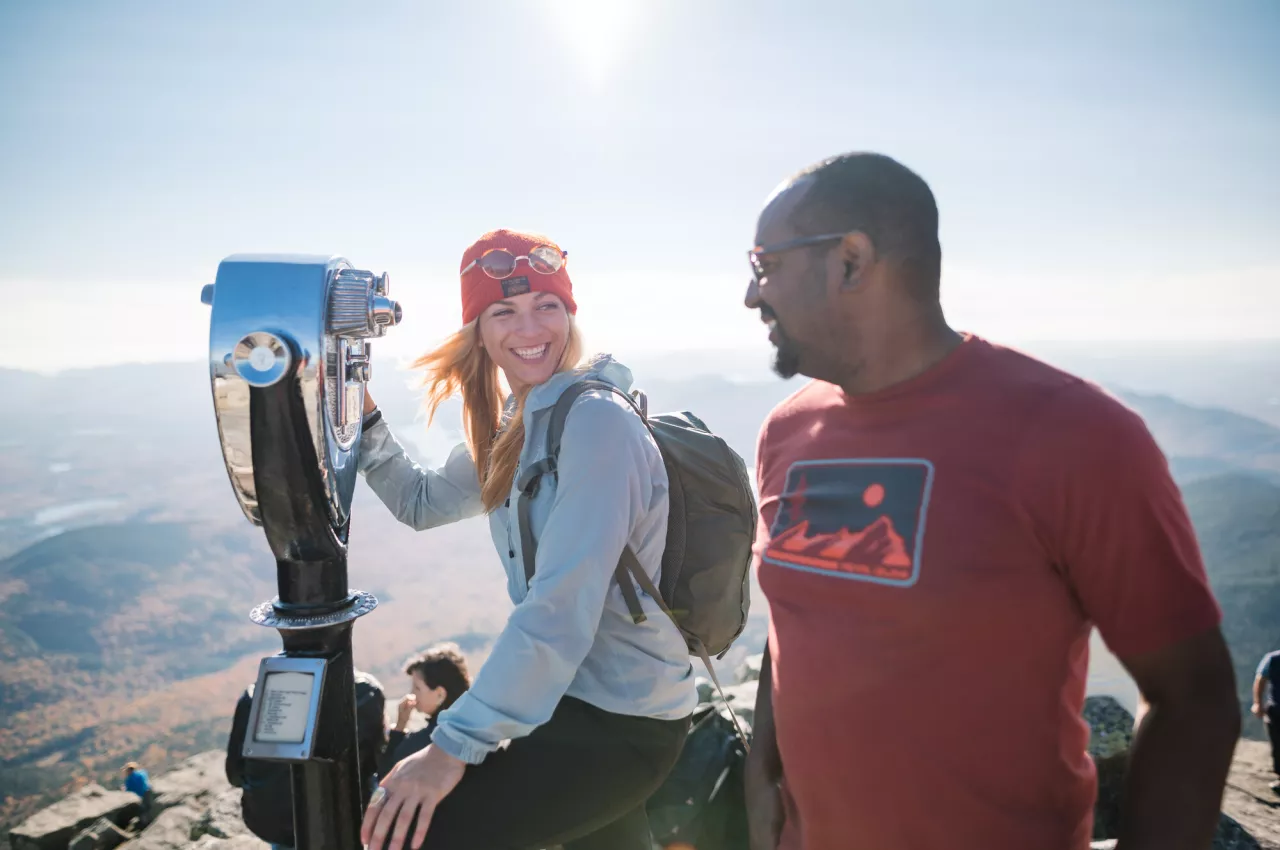 A woman and man look into a binocular stand on top of a mountain. 