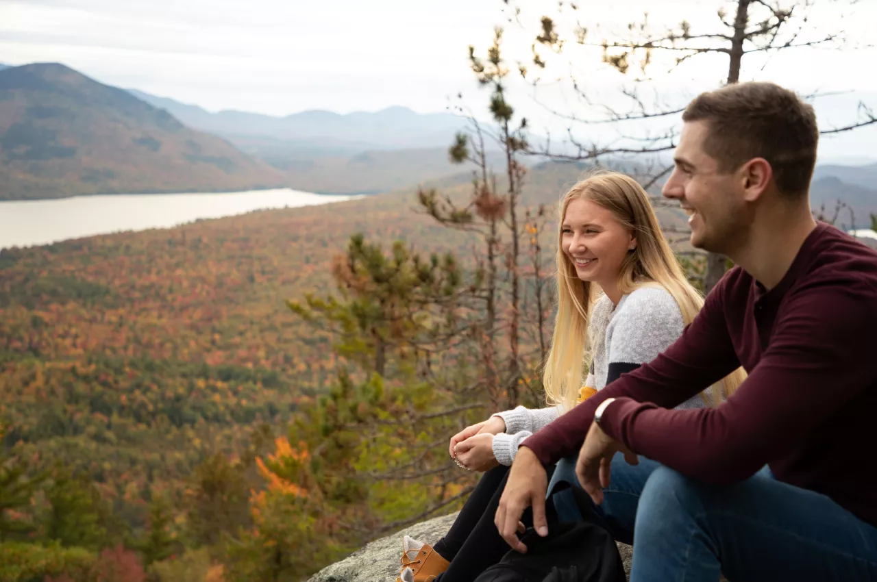 A man and woman sit on mountaintop looking at view of fall foliage, lake, and mountains