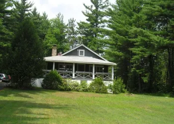 Exterior view of a cottage set back on a large green lawn with an ample covered porch and river stone chimney