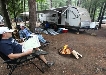 Men lounge in lawn chairs by a fire ring near an RV