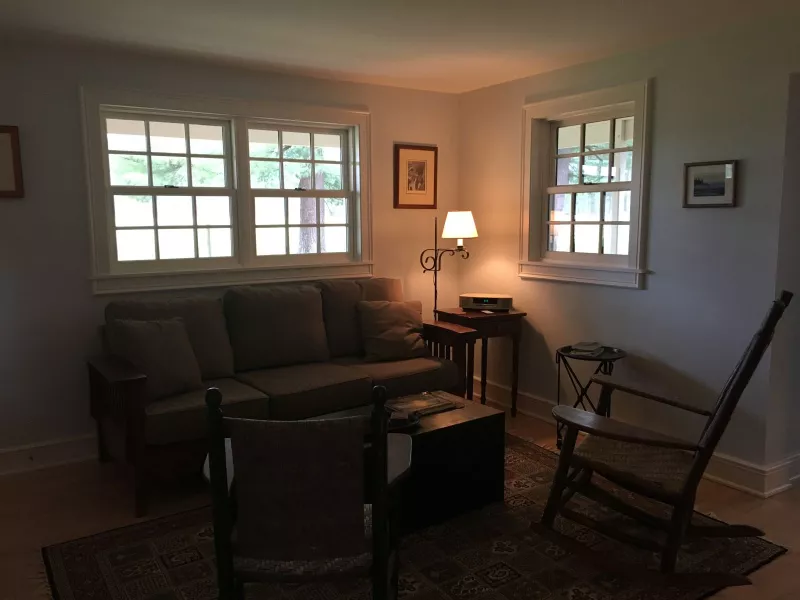A couch and two rocking chairs are in a small room dimly lit by a lamp and sunshine entering three window