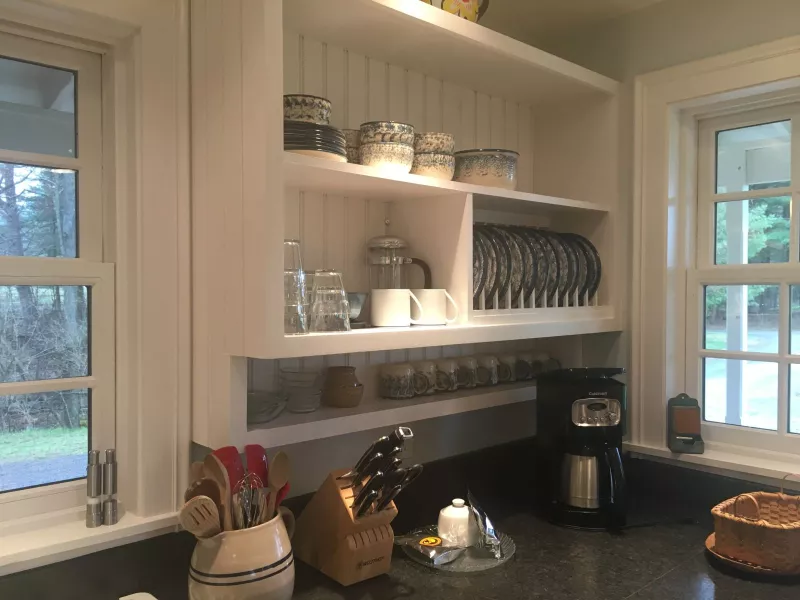 Plates, cups, mugs, and bowls are organized on open kitchen shelves with a coffee maker, knives, and cookware on the counter below