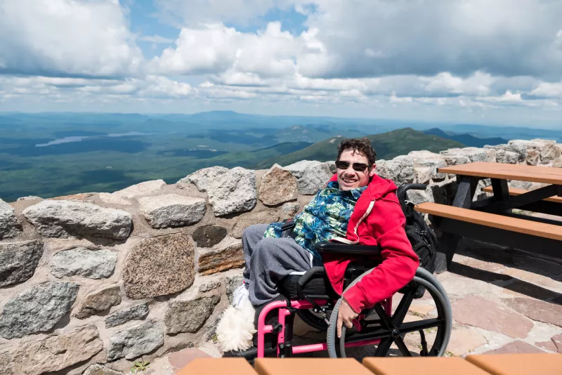 A man in a wheelchair poses next to a stone wall overlooking the Adirondacks below Whiteface Mountain.