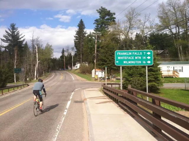 A cyclist passes a sign for Franklin Falls, Whiteface Mountain, and Wilmington