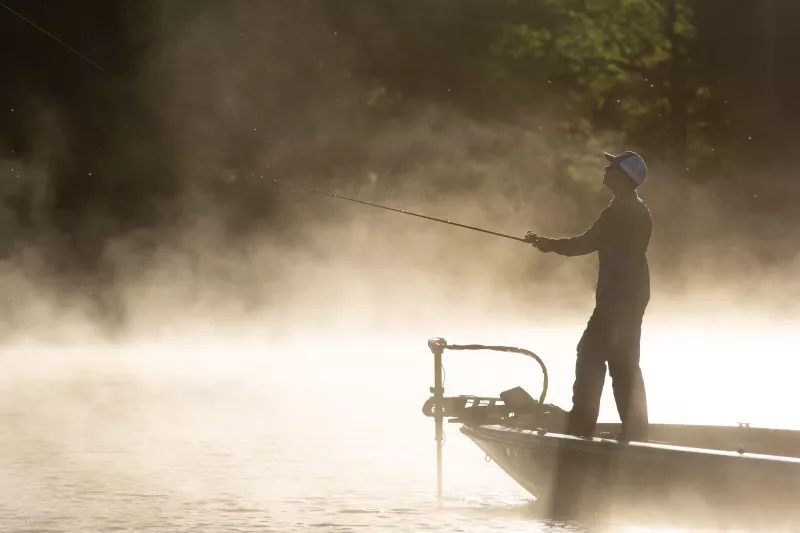 A man casts his line into the water while fishing on a boat in the fog.