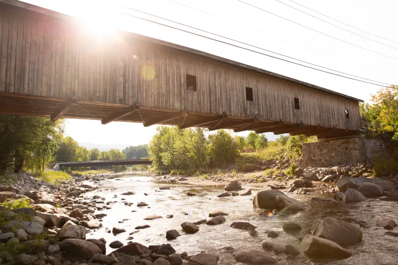 The sun shines over a historic covered bridge above a sparkling river filled with boulders.