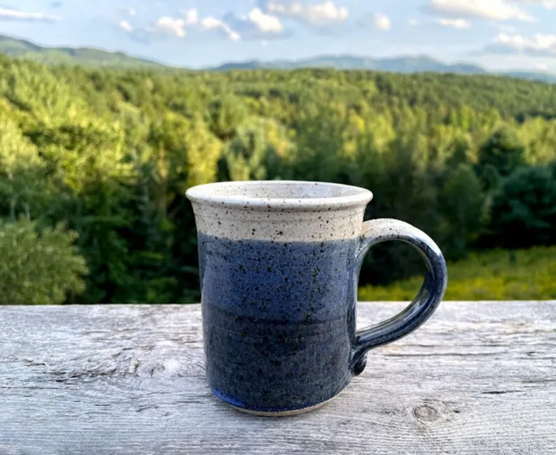A handmade pottery mug sits on a wooden railing overlooking a green mountain setting.