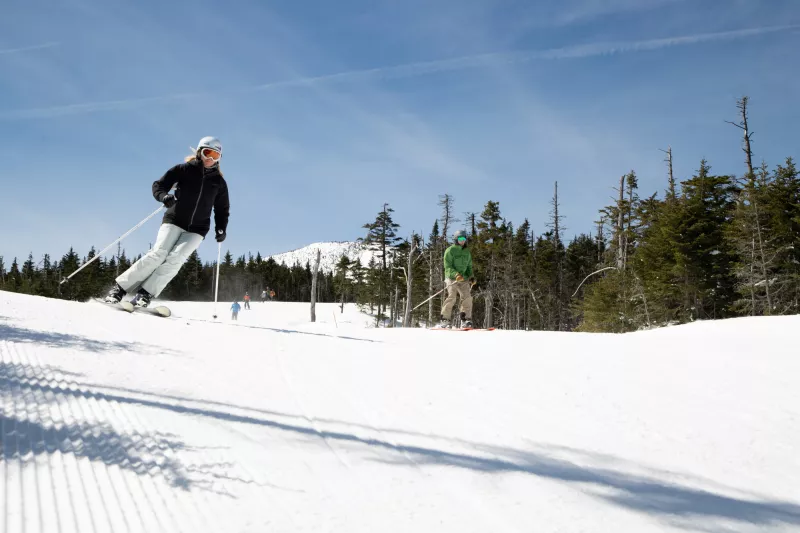 Skiers and snowboarders cruise down a groomed trail with Whiteface's summit in the background.