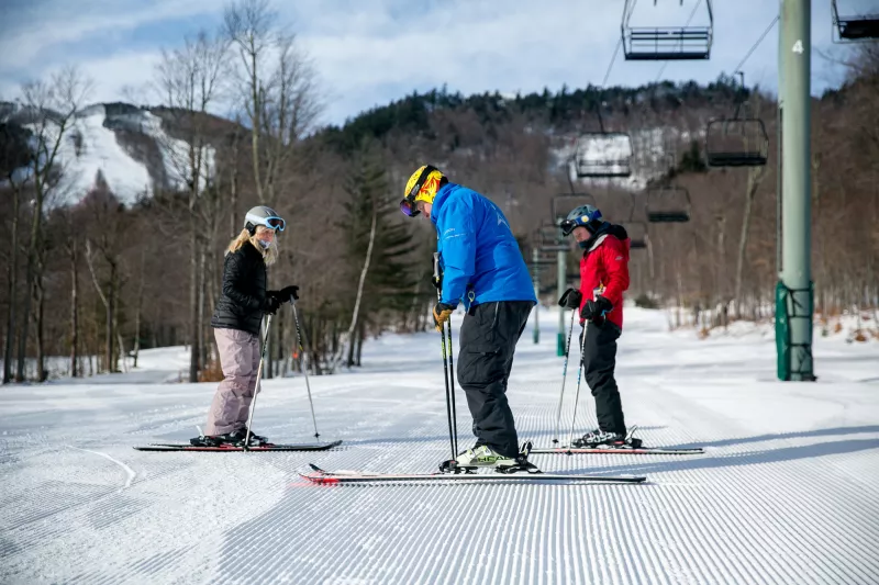 Two skiers with a ski instructor during a lesson on groomed trail near a chair lift.