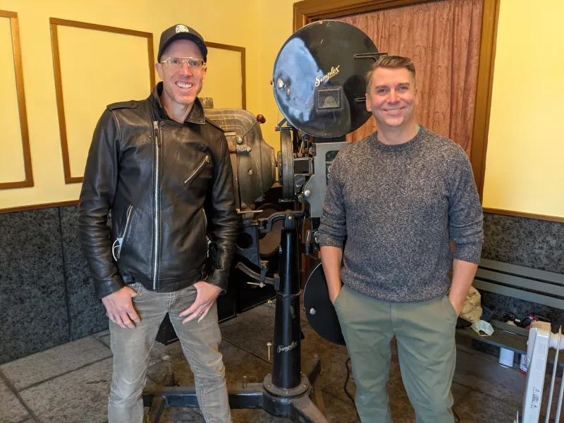 Two men pose in front of a large, old movie film projector