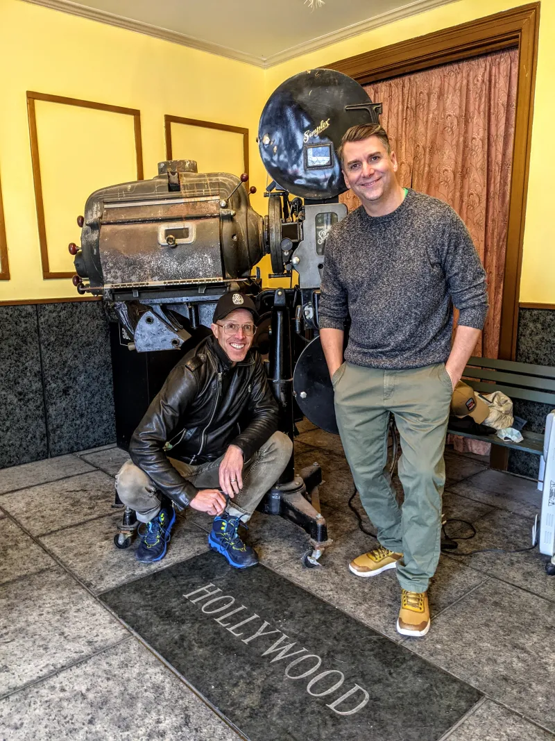 Two men pose in front of a large, old movie film projector